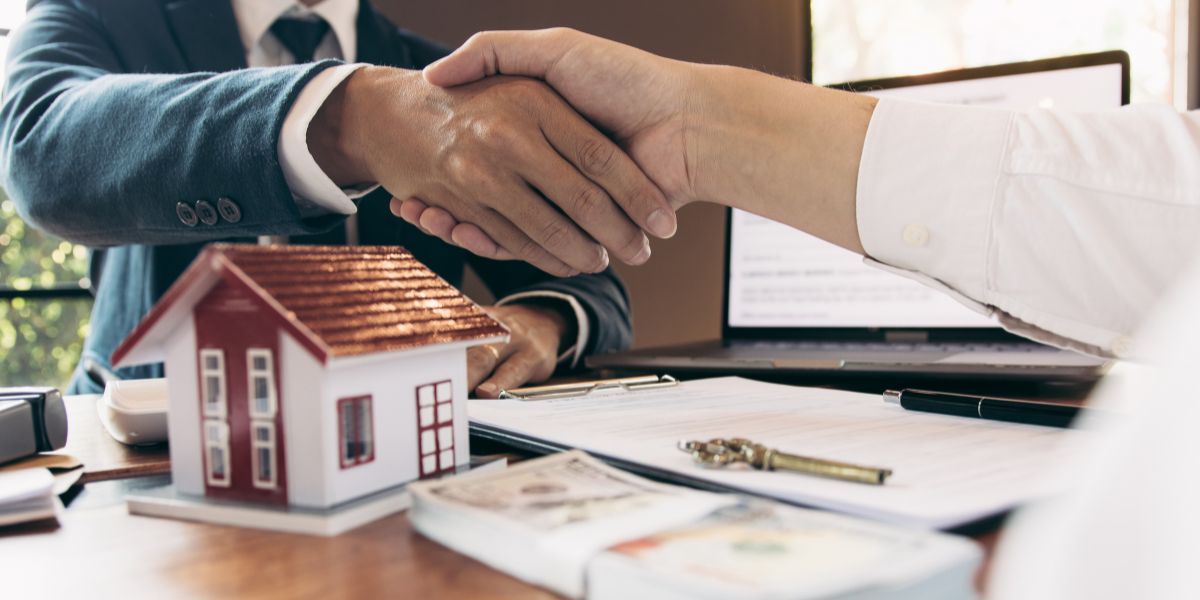 Two people shaking hands over a table with a house model, documents, and money, symbolizing a real estate deal.