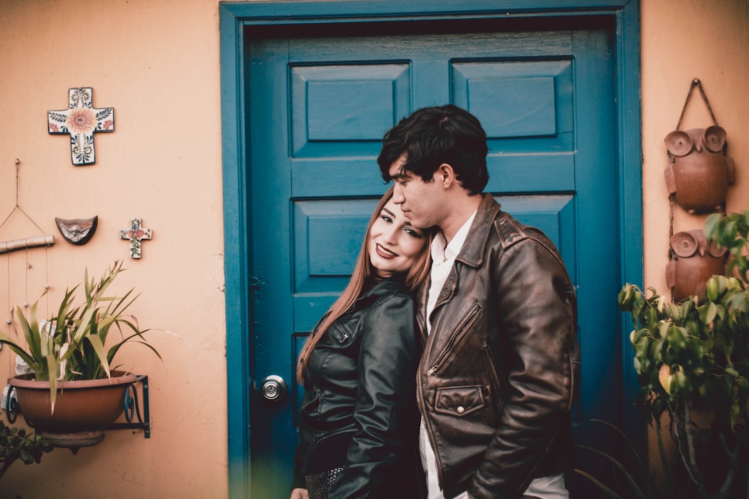A happy, stylish unmarried couple stands proudly in front of their new modern home, holding up the keys to the front door with smiles.