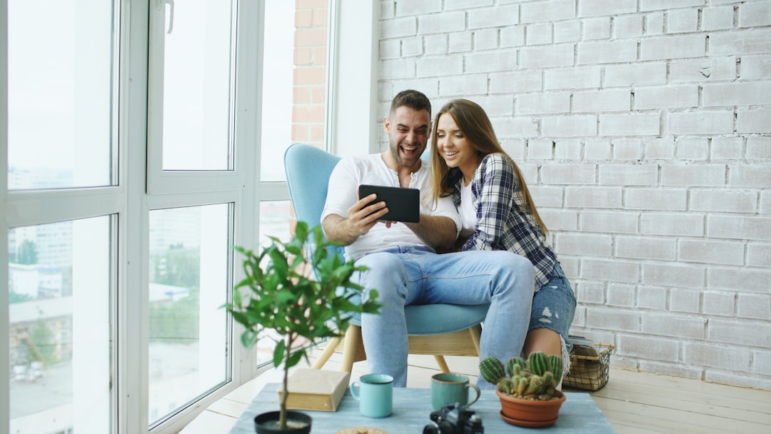 A young couple sits together on a sofa in a bright, modern living room, collaboratively using a laptop to plan their finances and home search.